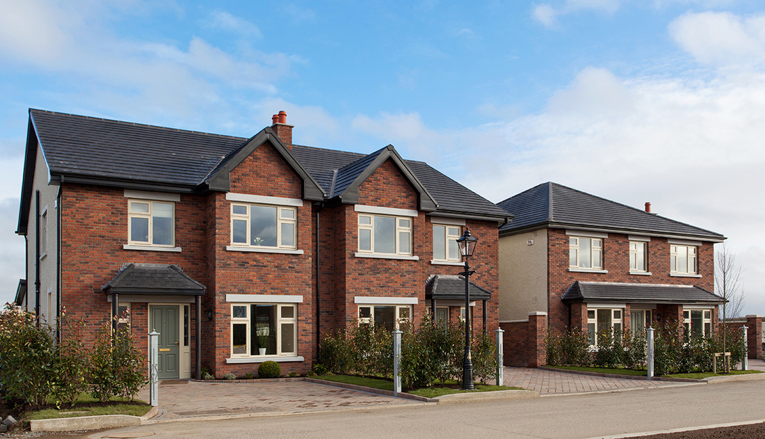 A two story, dark red, brick semi-detached house showcasing three windows. The driveway is laid with brick and there is lines of shrubs along the driveway. The driveway is made from brick and there is two blue posts on either side. There is two patches of grass with small trees on either side of the driveway.