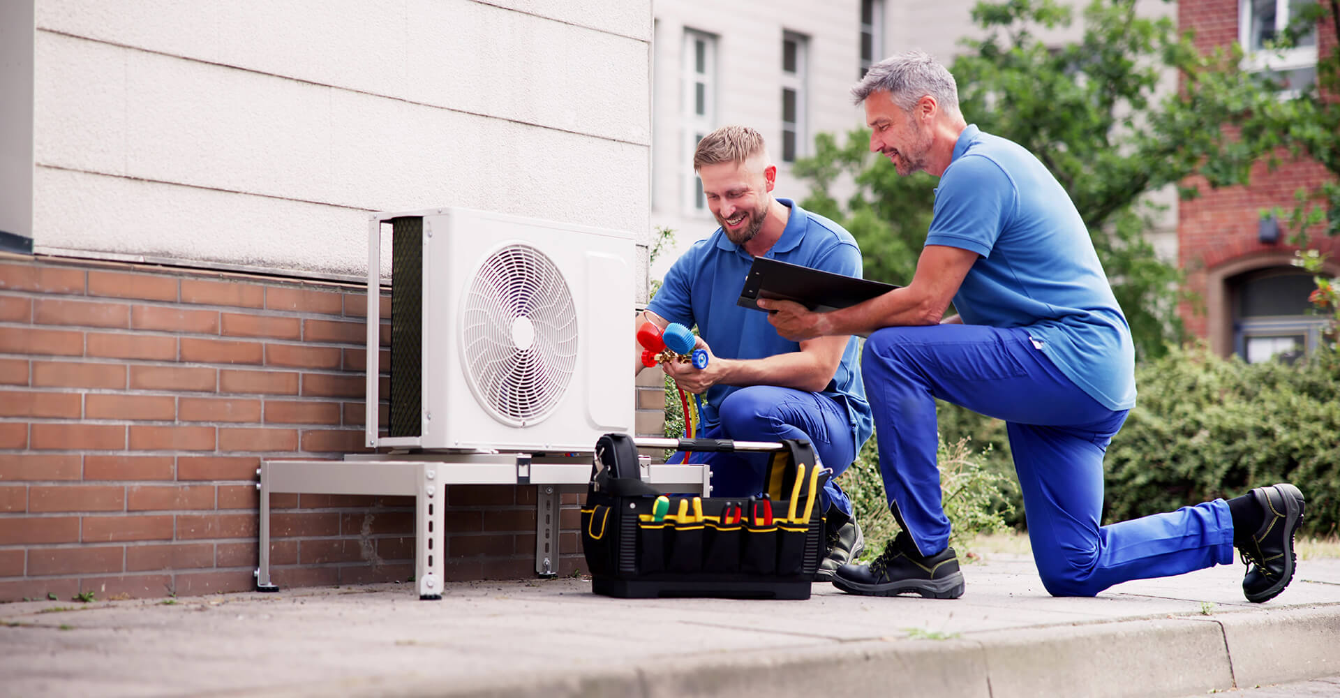 Two workers dressed in a blue uniform and black boots outside a building. They are knelt down beside a heater holding a red and blue tool. Beside them a tool box can be seen.