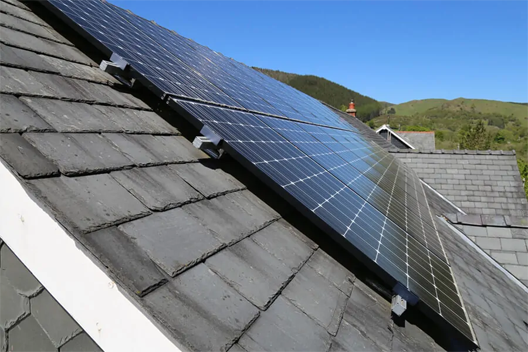 Solar panels built onto the slant of a tiled roof, reflecting the bright blue sky. Mountains can be seen in the background.