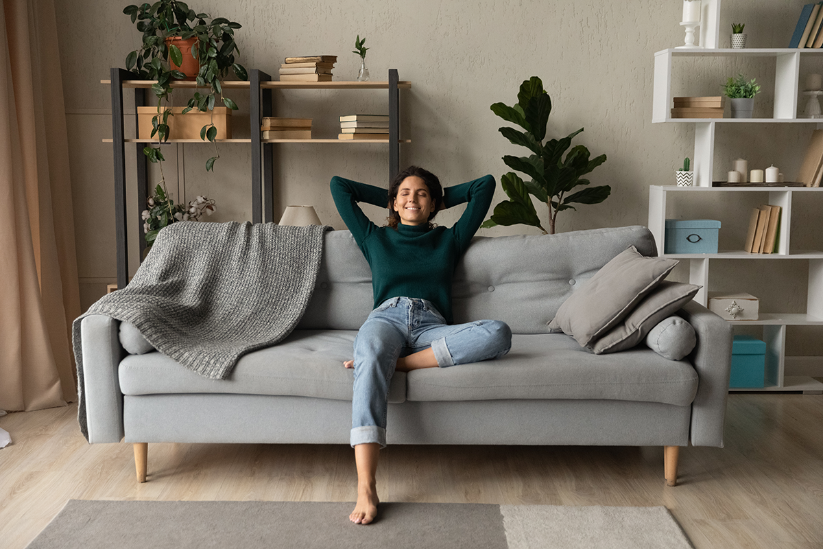A young woman in a green sweater and jeans relaxing on a modern gray couch with her hands behind her head, smiling with her eyes closed in a cozy, stylish living room.